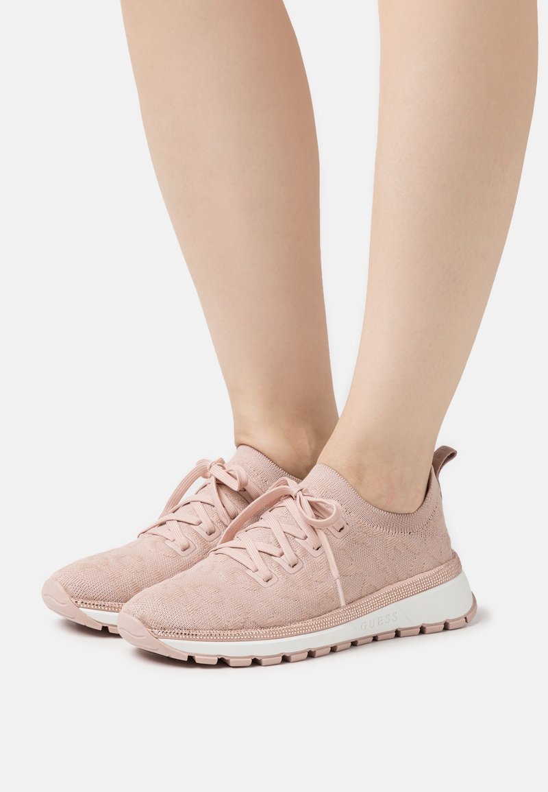 Pieds portant des baskets tricotées roses pâles avec des lacets et des semelles blanches, posés sur une surface blanche.