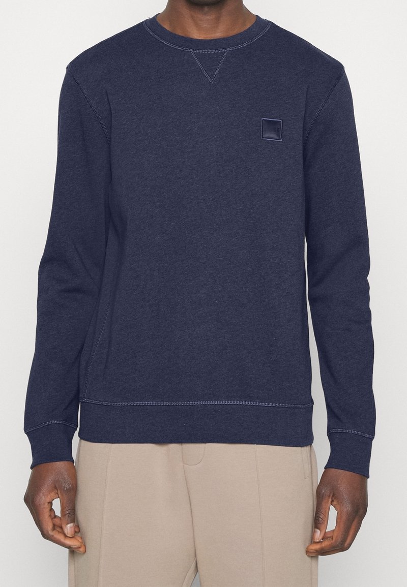 Homme portant un sweatshirt bleu marine à manches longues et col rond avec un petit logo carré sur la poitrine et un pantalon beige, debout devant un fond uni.