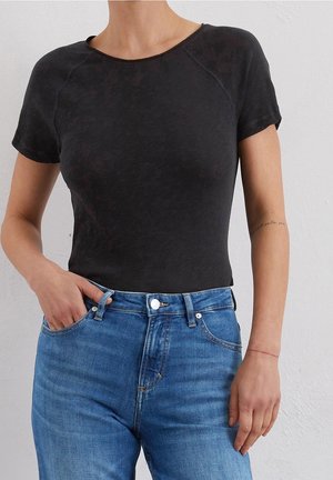 Kobieta w dopasowanym czarnym krótkim rękawem T-shircie, włożonym do wysokich niebieskich dżinsów, stoi z jedną ręką w kieszeni na tle białej ściany.