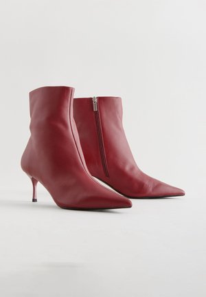 Bottes cheville en cuir rouge avec un bout pointu, un talon stiletto fin et une fermeture éclair latérale. Texture lisse avec une silhouette ajustée.
