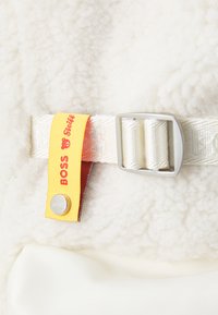 White fleece fabric with a yellow tag featuring "BOSS" and a bear logo. Silver hardware with a narrow strap and buckle detail. Soft texture.