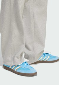 Light blue sneakers made of suede and mesh, featuring white and gold stripes, with a textured sole and prominent tongue detail.