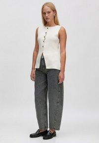 Weißes ärmelloses Oberteil mit Knopfleiste und subtiler Textur, kombiniert mit locker sitzenden dunkelgrauen Jeans und schwarzen Loafers.