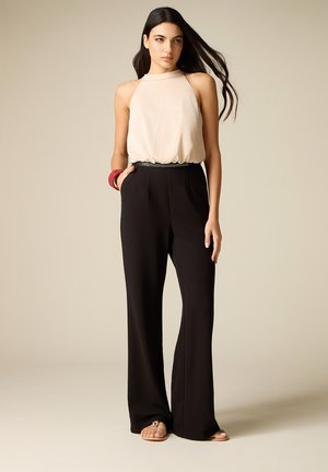 Femme portant un top beige sans manches, un pantalon noir large taille haute, un bracelet rouge et des sandales plates, debout devant un fond beige.