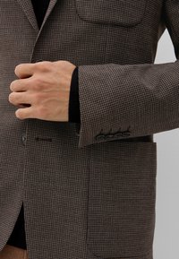 Braun kariertes Blazer mit strukturiertem Finish, ausgestattet mit einer Tasche, geknöpften Manschetten und einem Reverskragen. Der Stoff ist aus dunklen und hellen Fäden gewebt.