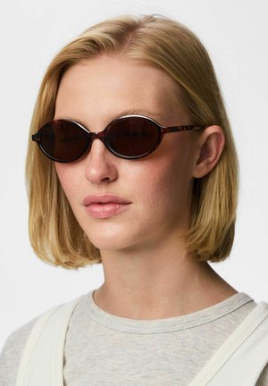 Joven con pelo rubio corto que lleva gafas de sol ovaladas con montura carey y una camiseta gris claro sobre un fondo blanco.