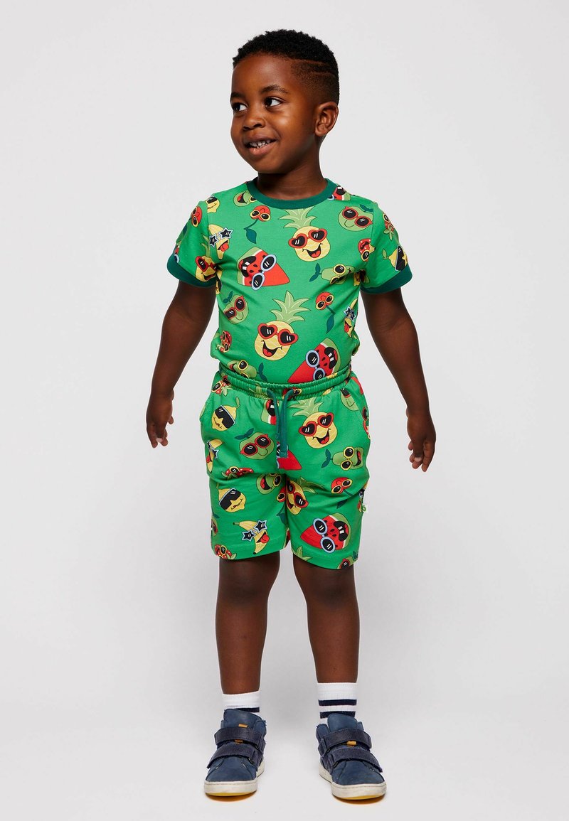 Jeune garçon portant une chemise et un short verts assortis ornés d'ananas et de pastèques colorés et dessinés, debout et souriant.