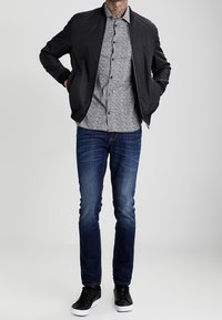 Blouson aviateur noir porté sur une chemise à manches courtes à motifs, associé à un jean bleu et des baskets noires. Tissu lisse et silhouette moderne.