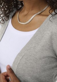 Cardigan gris avec un col en V superposé à une chemise blanche ajustée. Le cardigan présente une texture douce. Un collier chaîne en argent est porté.