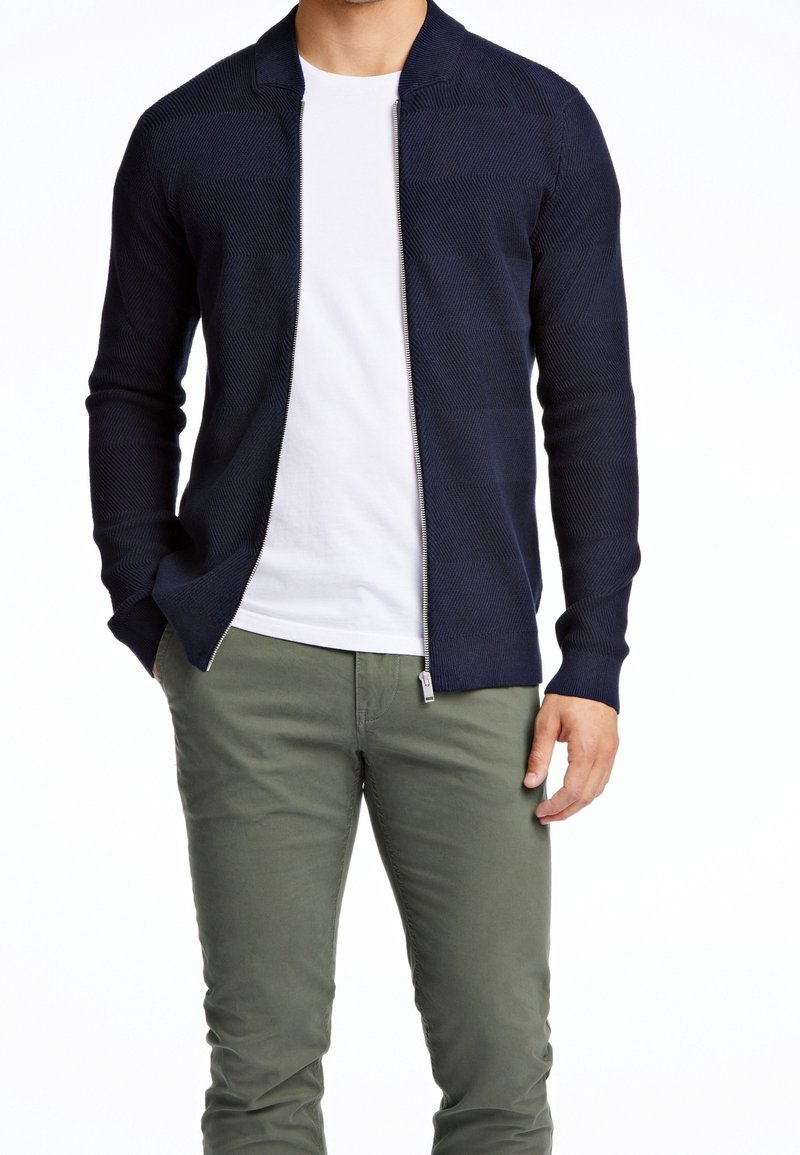 Homme portant un cardigan zippé bleu marine sur un t-shirt blanc avec un pantalon vert olive, debout, une main dans la poche.