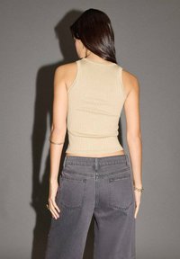 Žena s dlouhými tmavými vlasy, oblečená v bezrukávovém béžovém žebrovaném topu a šedých džínech s vysokým pasem, stojící na pozadí v uniformní šedé barvě.