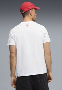Weißes Baumwoll-T-Shirt mit rundem Ausschnitt, kurzen Ärmeln und einem kleinen roten Logo im oberen Rückenbereich. Getragen mit dunklen Jeansshorts.