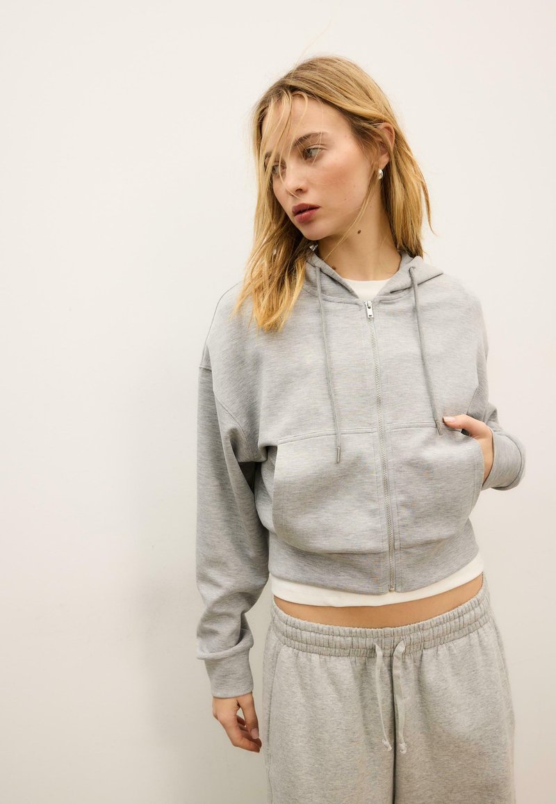 Sweat à capuche gris raccourci avec une fermeture éclair avant, un cordon de serrage et deux poches avant, associé à un pantalon de survêtement gris avec une taille élastique.