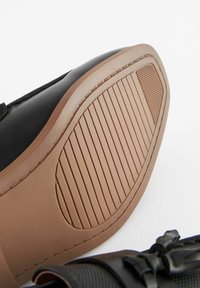 Chaussure noire avec un dessus lisse, dotée d'une semelle en caoutchouc beige. La semelle présente des stries pour l'adhérence et un motif texturé sur le côté.