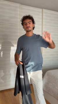 Άνδρας με σγουρά μαλλιά που φοράει μπλε πουκάμισο και άσπρο παντελόνι κρατάει ένα μαύρο σακάκι σε ένα χέρι και κουνάει το άλλο σε ένα υπνοδωμάτιο.