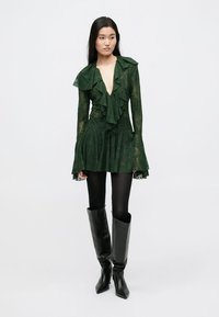 Πράσινο μακρυμάνικο φόρεμα από δαντέλα με γιακά και μανίκια με βολάν, καμπυλωτό τελείωμα, συνδυασμένο με μαύρες μπότες που φτάνουν μέχρι τους μηρούς και μαύρο καλσόν.