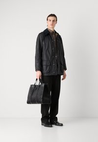 Veste noire imperméable avec un col bleu contrastant, chemise marron en dessous, pantalon noir et sac fourre-tout noir texturé.