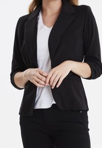 Svart kavaj gjord av slät tyg, med enknapponknäppning, spetsiga slag och långa ärmar, bärs över en vit T-shirt.