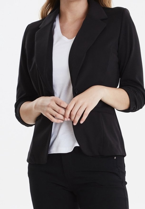 Svart kavaj gjord av slät tyg, med enknapponknäppning, spetsiga slag och långa ärmar, bärs över en vit T-shirt.