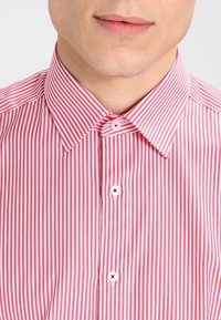 Camicia a righe rosse e bianche con colletto a punta, dotata di bottoni bianchi con dettagli neri e una texture di tessuto liscia.