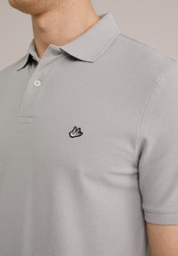 Polo de culoare gri deschis, realizat dintr-un material texturat. Dispune de un guler clasic și un mic logo brodat, de culoare neagră, pe pieptul stâng.