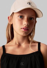 Top negro sin mangas con textura arrugada y un pequeño logo "CK". Gorra de béisbol beige claro con un "CK" bordado en la parte frontal.