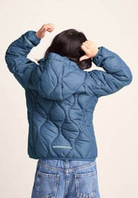 Child in blue quilted jacket with hood raised arms, wearing blue denim jeans, facing away against a plain background.