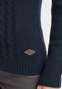 Dunkelblauer gestrickter Pullover mit einem Zopfmuster auf der Vorderseite und einem gerippten Saum. Enthält ein braunes, lederähnliches Logopatch.