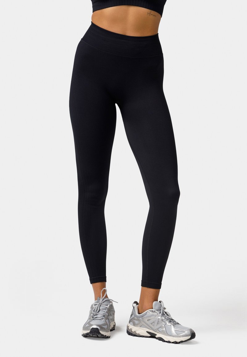 Schwarze Leggings mit hohem Bund, die eine gerippte Textur und nahtloses Design aufweisen. Kombiniert mit silbernen Sportschuhen.