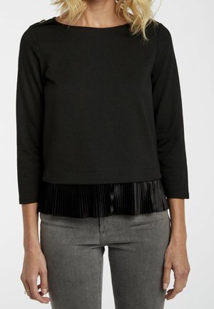 Femme portant un haut noir à manches longues avec un détail plissé à l'ourlet et un jean gris, debout devant un fond clair uni.
