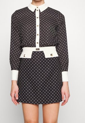 Czarna sukienka w wzory z białym kołnierzykiem, mankietami i paskiem w talii, ozdobiona złotymi ćwiekami w kształcie kwadratów na guzikach i akcentach paska.