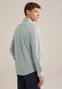 Tânăr cu păr scurt și creț, îmbrăcat cu o cămașă lungă de culoare albastră deschisă și pantaloni închiși, stând și privind spre dreapta.