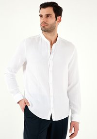 Camisa blanca de botonadura con tejido texturizado, cuello ajustado y mangas largas. Presenta un dobladillo curvado y un ligero brillo.