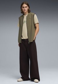 Veste sans manches vert olive avec une fermeture éclair à l'avant, portée sur un t-shirt beige clair ; associée à un pantalon large foncé et des chaussures beiges.