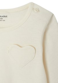 Sahnfarbener Baby-Overall mit einem kleinen Herzpatch auf der Brust und einer Druckknopfverschlus an der Schulternaht.