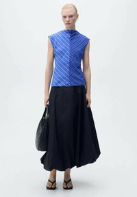 Blue, sleeveless top with diagonal stripes paired with a long black skirt, carrying a black handbag; flat strapped sandals on feet.
