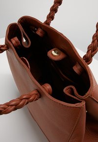 Bruine leren handtas met gevlochten handvatten, open bovenkant die een glad interieur onthult, en een gouden drukknopsluiting. Twee binnenzakken zichtbaar.