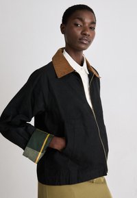 Casaco preto com um colarinho de veludo cotelê castanho, punhos com detalhes em verde e amarelo, fecho de zíper e bolsos laterais, apresentando um ajuste descontraído.