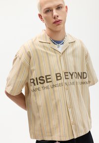 Rövid ujjú, bézs gombos ing, függőleges fehér csíkokkal és elülső "RISE BEYOND" felirattal. Pamut anyag, laza fazon.