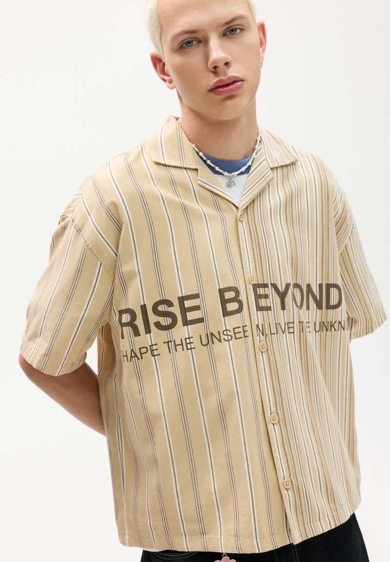 Rövid ujjú, bézs gombos ing, függőleges fehér csíkokkal és elülső "RISE BEYOND" felirattal. Pamut anyag, laza fazon.