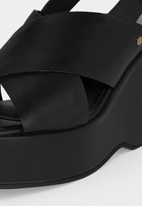 Μαύρο σανδάλι πλατφόρμας με φαρδιές σταυρωτές δερμάτινες λωρίδες και χρυσή λεπτομέρεια με πριτσίνι στο πλάι.