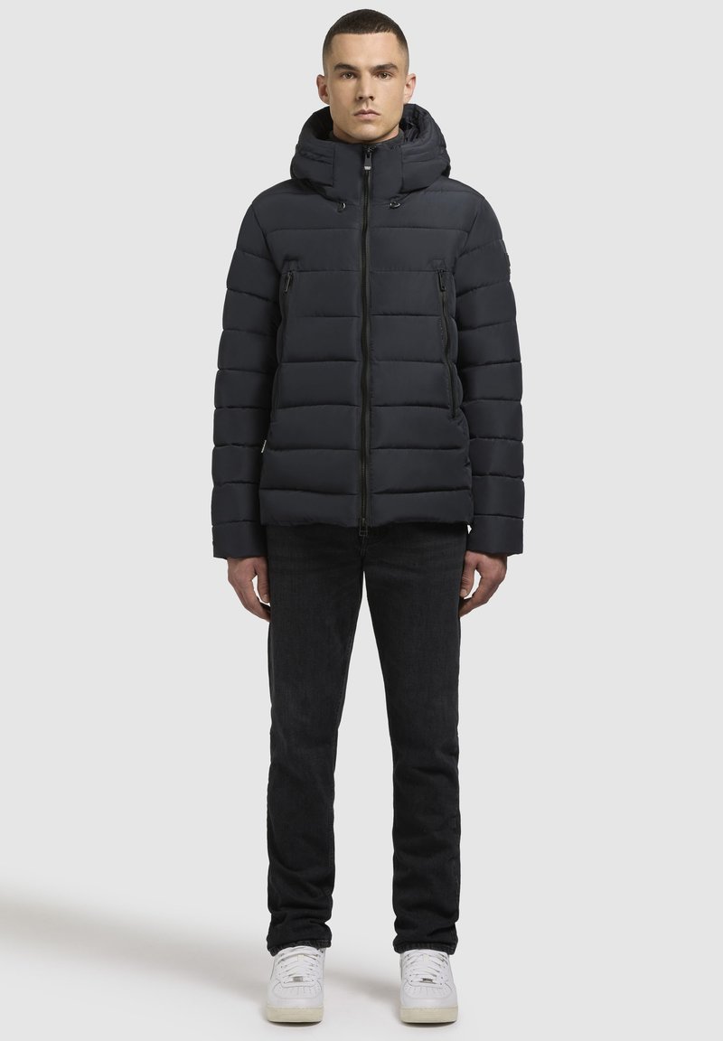 En vuxen man står vänd framåt, klädd i en svart puffjacka, svarta jeans och vita sneakers mot en enfärgad ljus bakgrund.