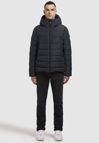 Sort pufferjakke med vandret syninger, lynlåslukning og hætte. Bæres over mørke jeans og hvide sneakers. Glat, polstret tekstur.