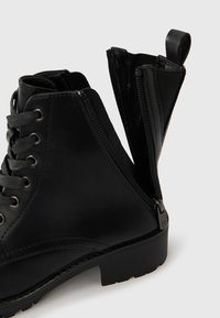 Schwarze Knöchelstiefel aus glattem Leder, mit runder Spitze, Schnürvorderseite und seitlichem Reißverschluss. Texturierte Sohle für besseren Halt.