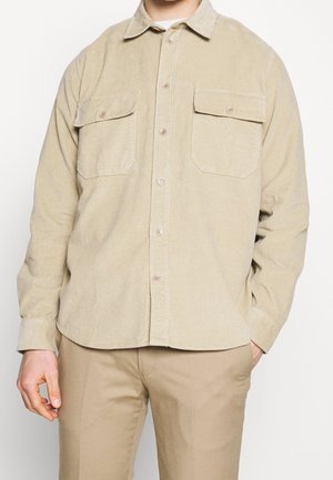 Mann, der ein beiges Cordhemd mit Brusttaschen und passende beige Hose trägt, steht vor einfachem Hintergrund.