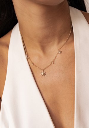 Gold chain necklace with sun, moon, and star charms worn on a person in a white sleeveless top.