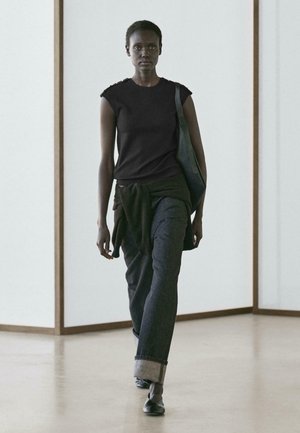 Donna che cammina al chiuso indossando una canotta nera senza maniche, jeans scuri con risvolti, ballerine nere e portando una grande borsa a tracolla nera.