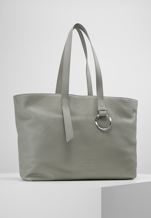 Sac cabas en cuir texturé gris avec deux longues poignées et un détail de bague argentée sur une poignée, reposant sur une surface blanche.