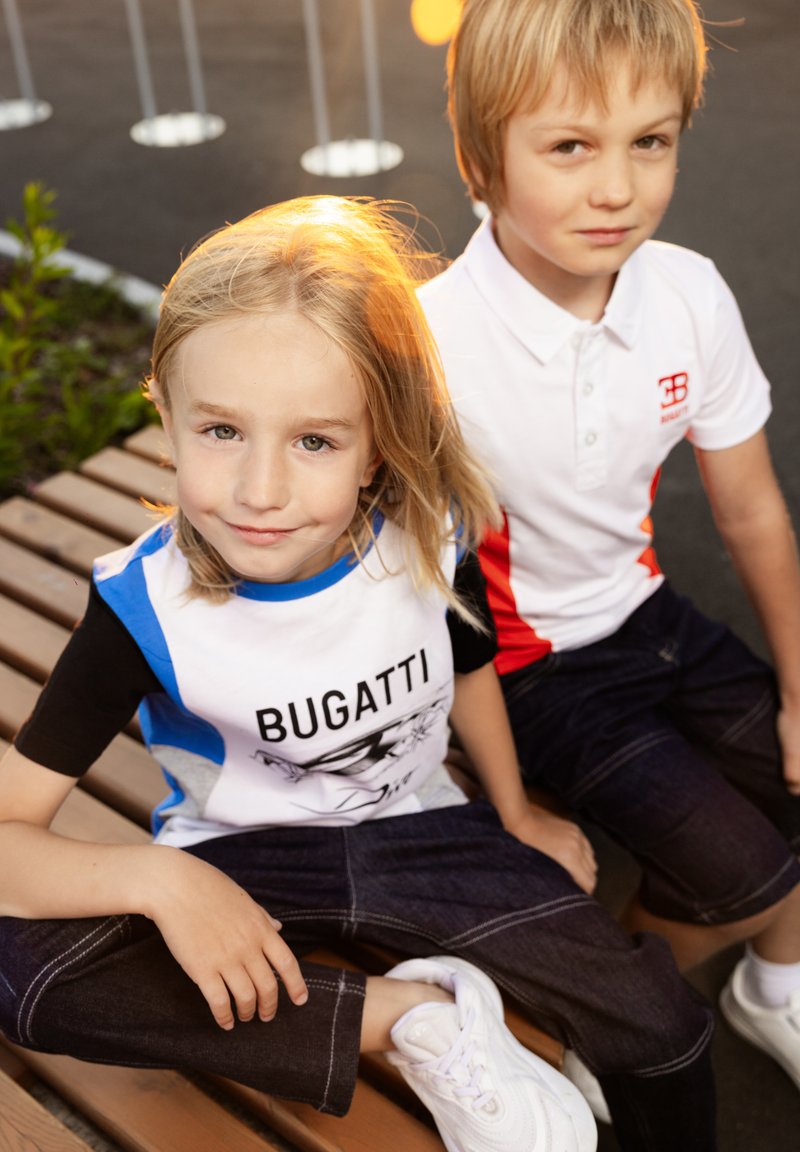 Due bambini sono seduti su una panchina di legno. Uno indossa una maglietta Bugatti blu, nera e bianca; l'altro ha un polo bianca con dettagli rossi. Entrambi indossano jeans scuri.