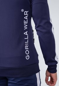 Marineblauwe sweatshirt met witte tekst "GORILLA WEAR" op de linkermouw, textiel met structuur, aangesloten ontwerp en ritssluitingsdetail in de zak.